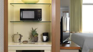 A hotel room with a kitchenette area featuring a microwave, a coffee maker, a small plant, and a glass container. There is a bed with a white comforter and a television on a wooden table.