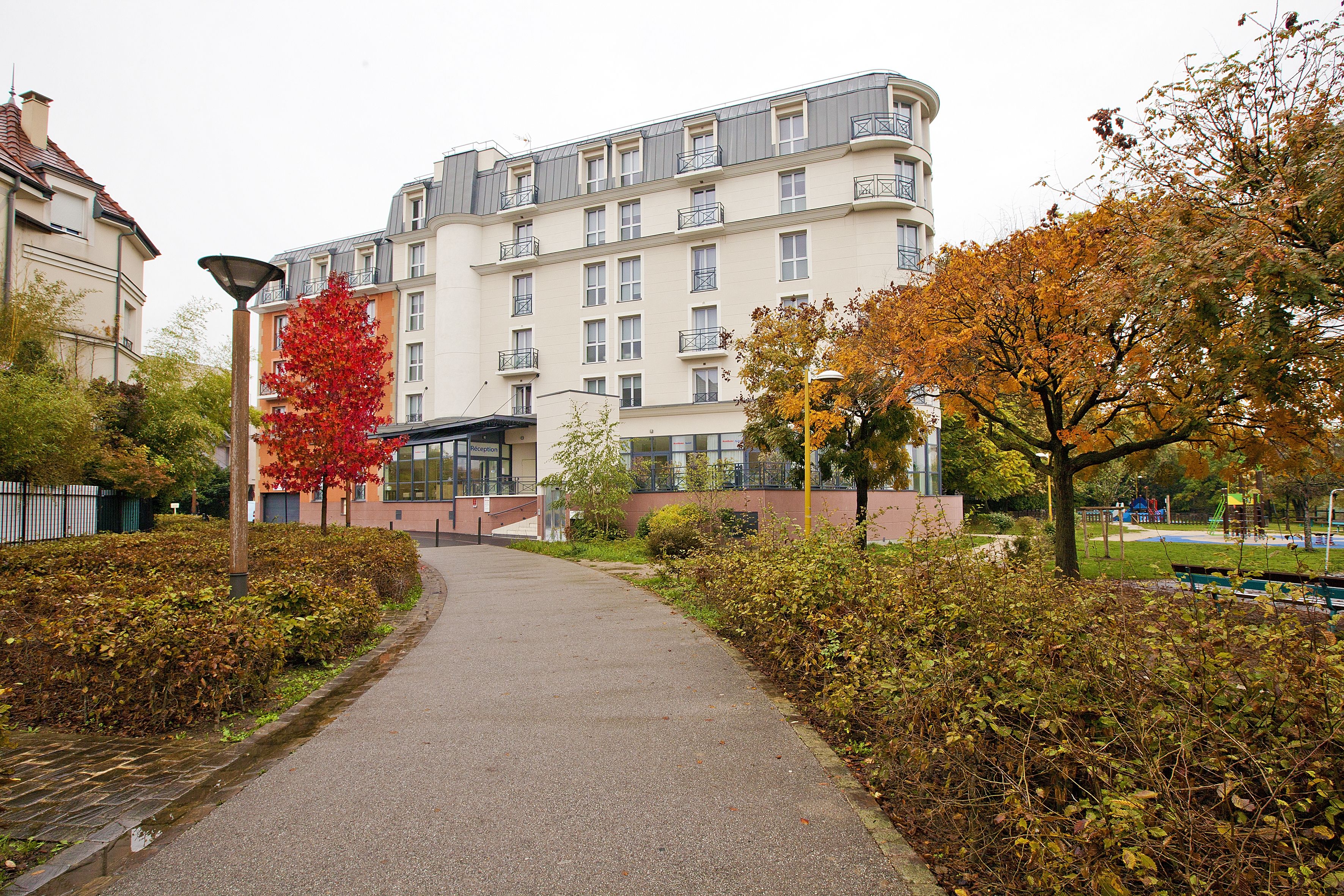 Un chemin menant à un immeuble à plusieurs étages doté de balcons, entouré d’un feuillage automnal et d’un espace vert aménagé en parc.