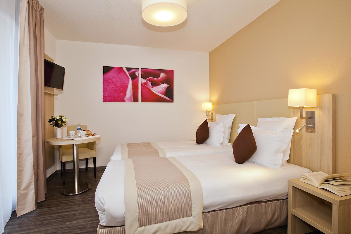 Een hotelkamer met twee bedden, een nachtkastje met lamp, een tafeltje met een bloevas en een flatscreen televisie aan de muur.