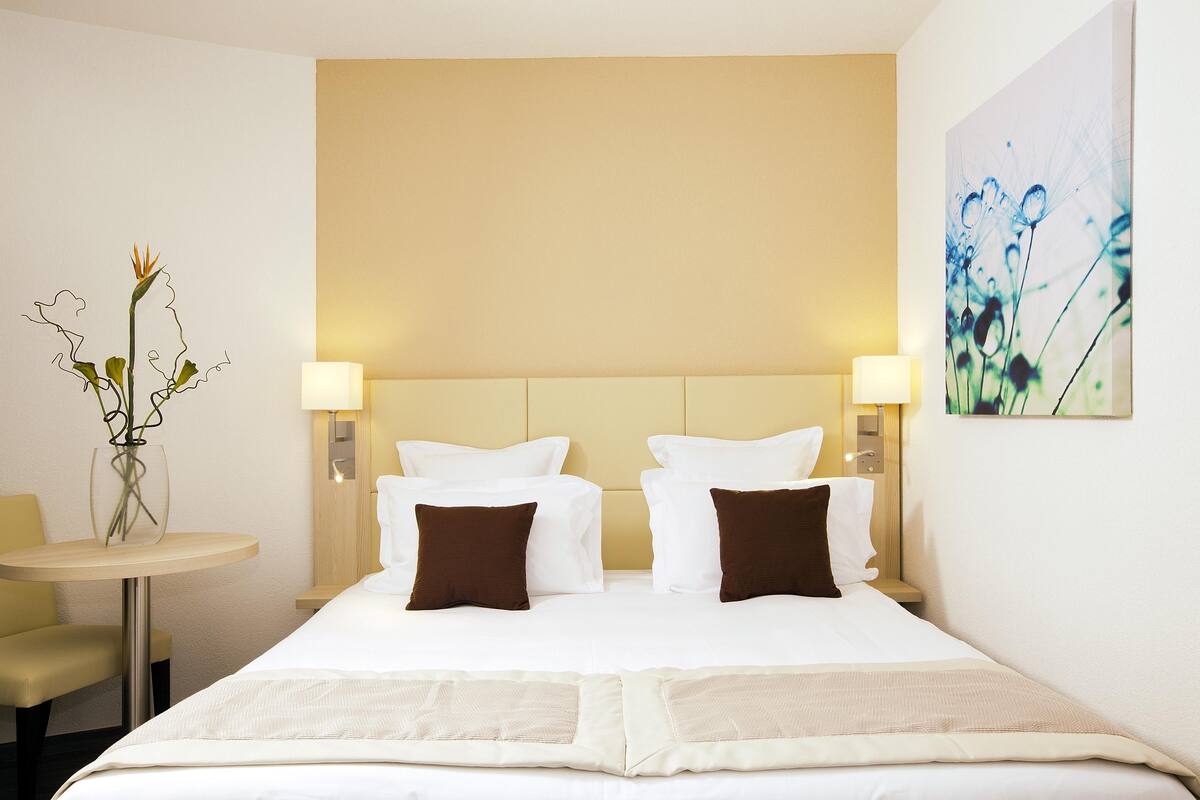 Een hotelkamer met een groot bed, twee nachtkastjes met lampen, een kleine ronde tafel met een vaas en een aan de muur hangend schilderij.