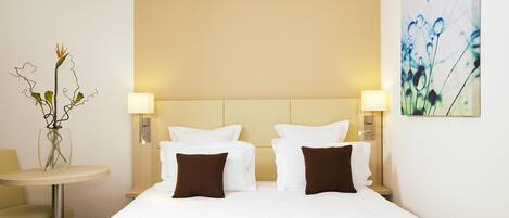 A hotel room with a large bed, two bedside tables with lamps, a small round table with a vase, and a wall-mounted painting.
