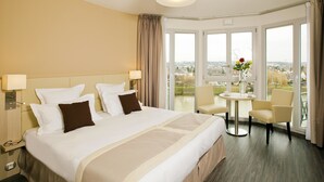A hotel room with a large bed, two chairs, a small table, and a view of the city through large windows.