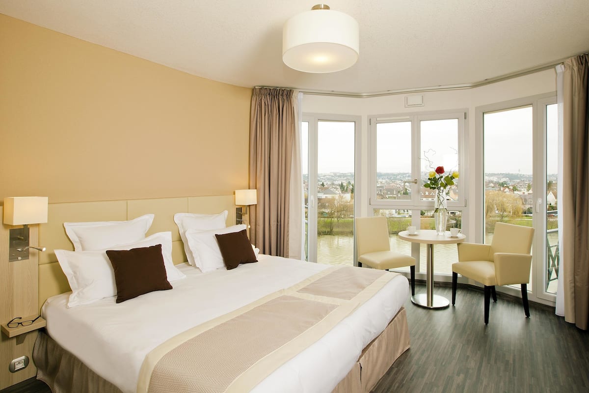 Hotelkamer met een groot bed, twee nachtkastjes, een ronde tafel met stoelen en uitzicht op de stad door grote ramen.