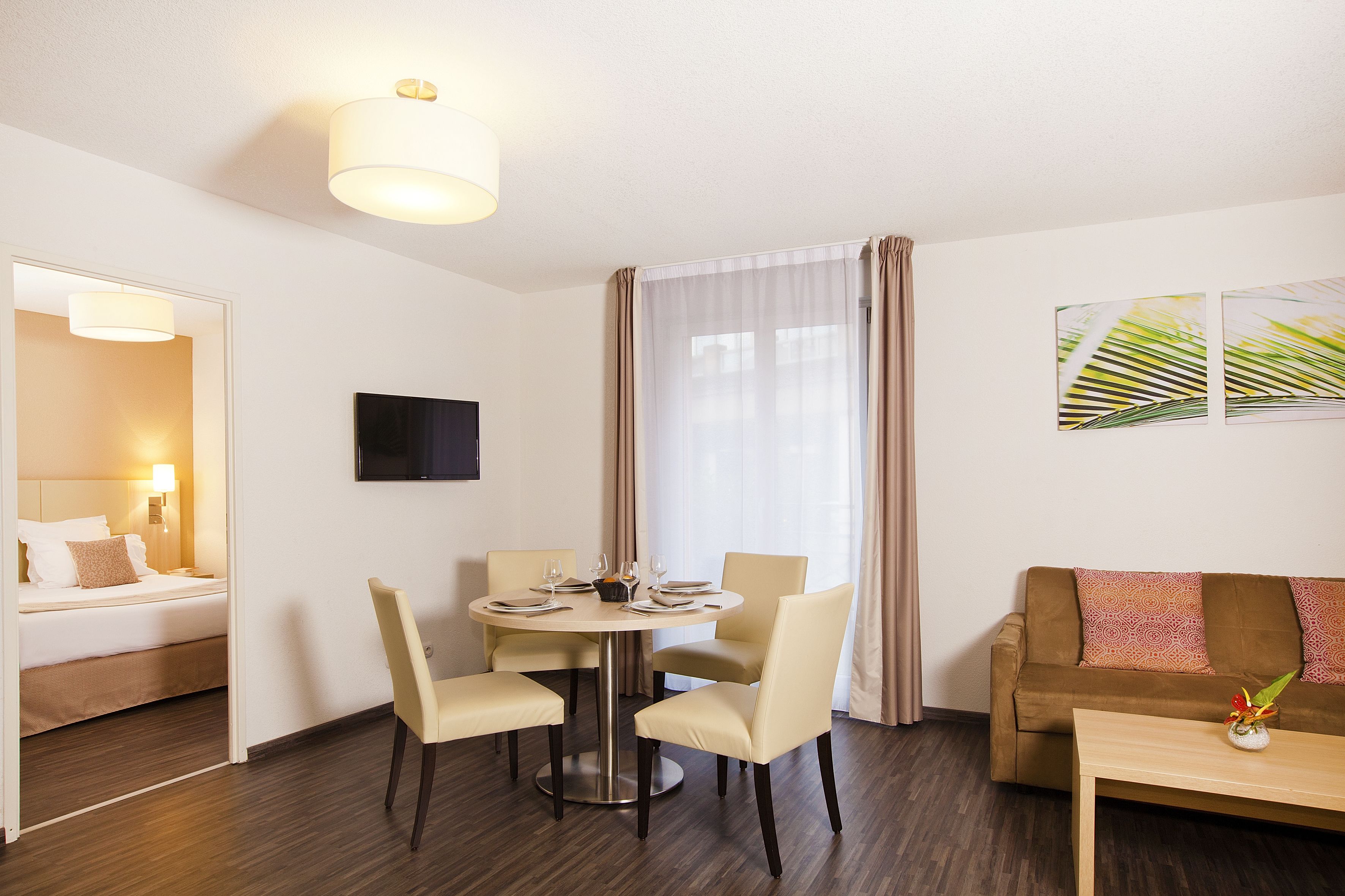 A hotel room with a bed, a round dining table with chairs, a sofa, and a TV mounted on the wall.