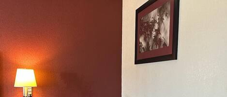 A room with a desk, chair, lamp, and a framed picture on the wall.