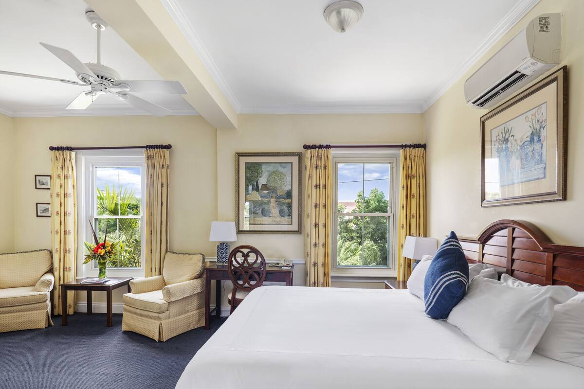 A hotel room with a bed, two armchairs, a desk, a ceiling fan, and large windows with curtains.