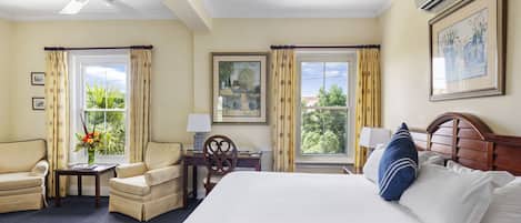 A hotel room with a bed, two armchairs, a desk, a ceiling fan, and large windows with curtains.