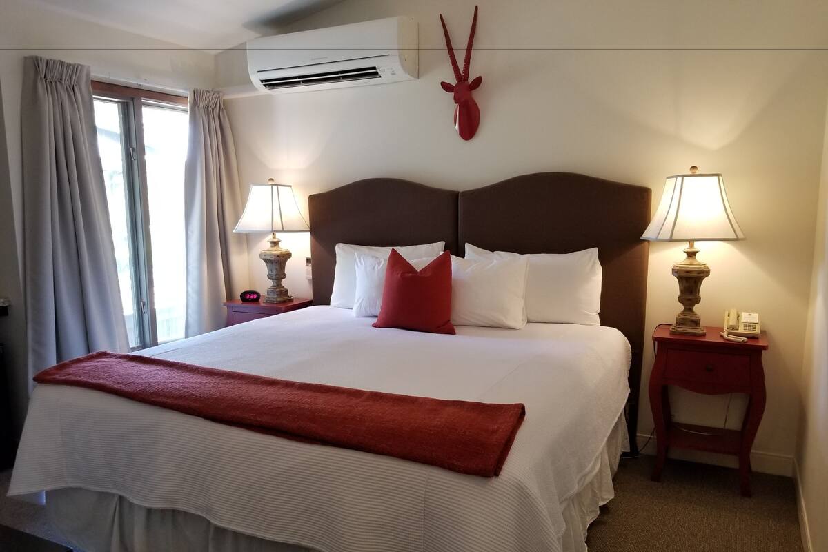 Cama bem arrumada com roupa de cama branca e vermelha, duas luminárias de cabeceira, um ar condicionado fixado na parede e uma cabeça de antílope decorativa.