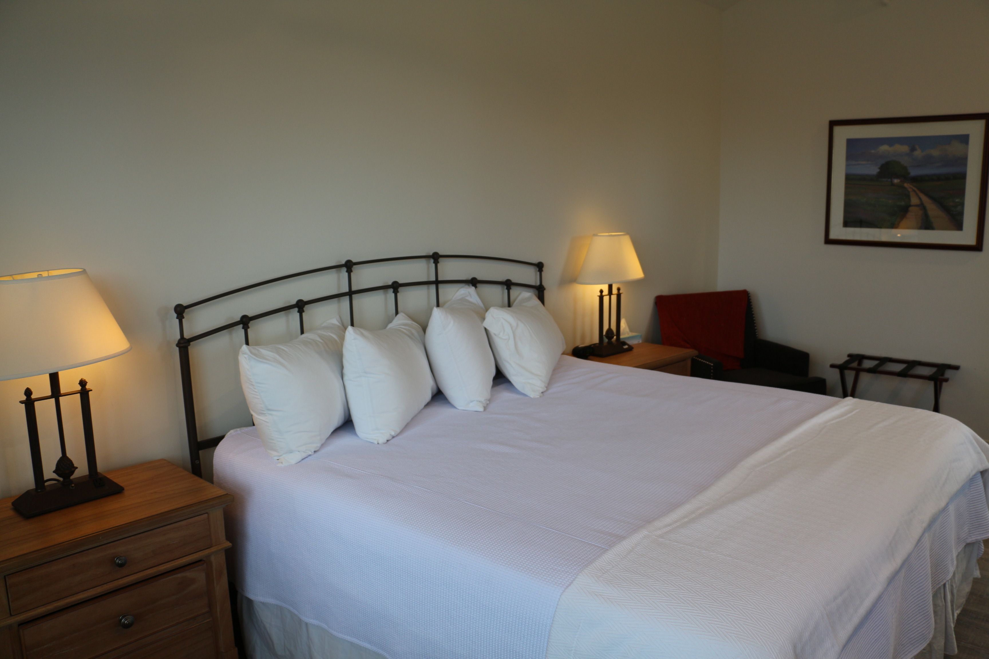 A neatly made bed with white linens and pillows, two bedside tables with lamps, a small red chair, and a framed picture on the wall.