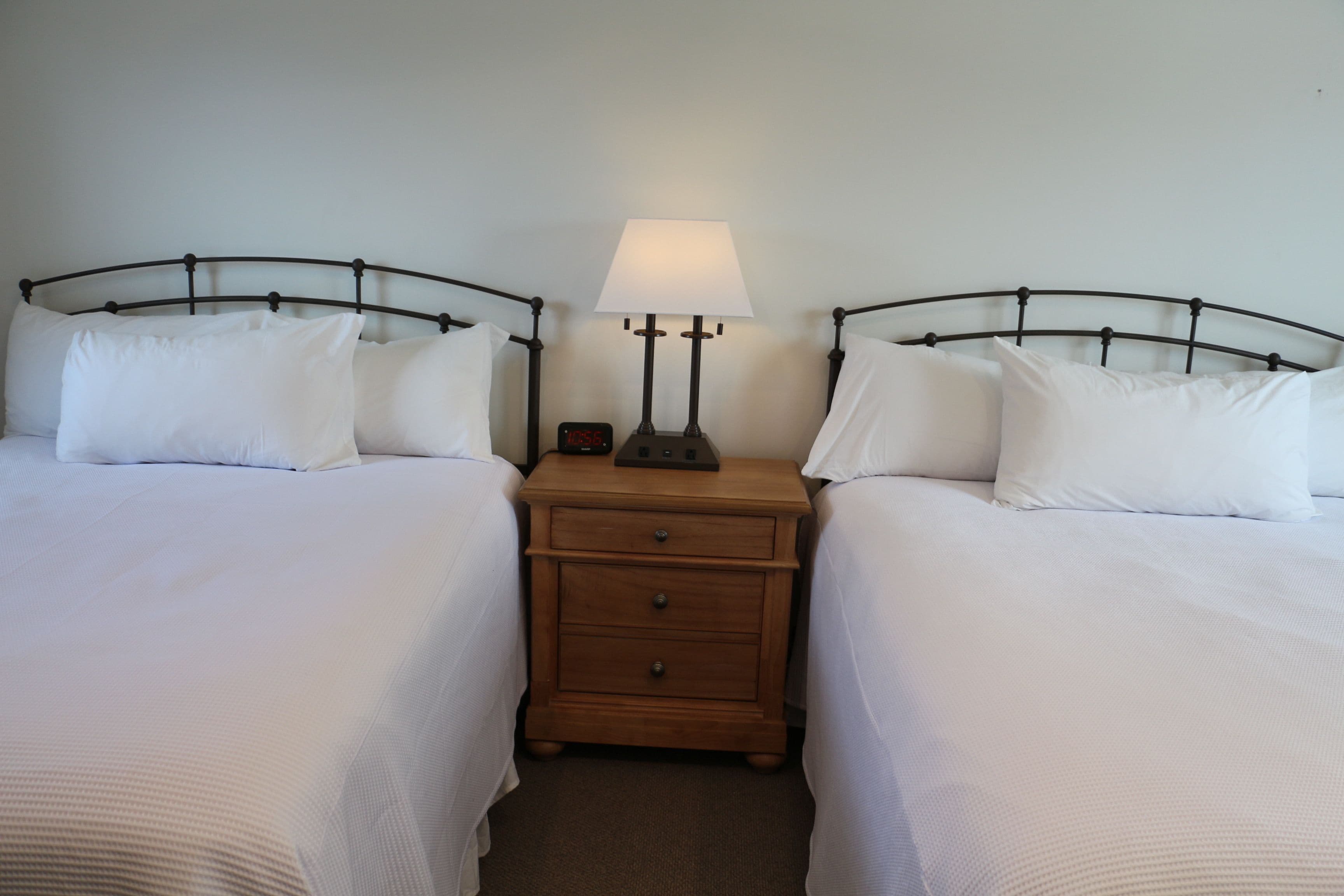 A hotel room with two single beds, a wooden nightstand with a lamp, and a digital clock.