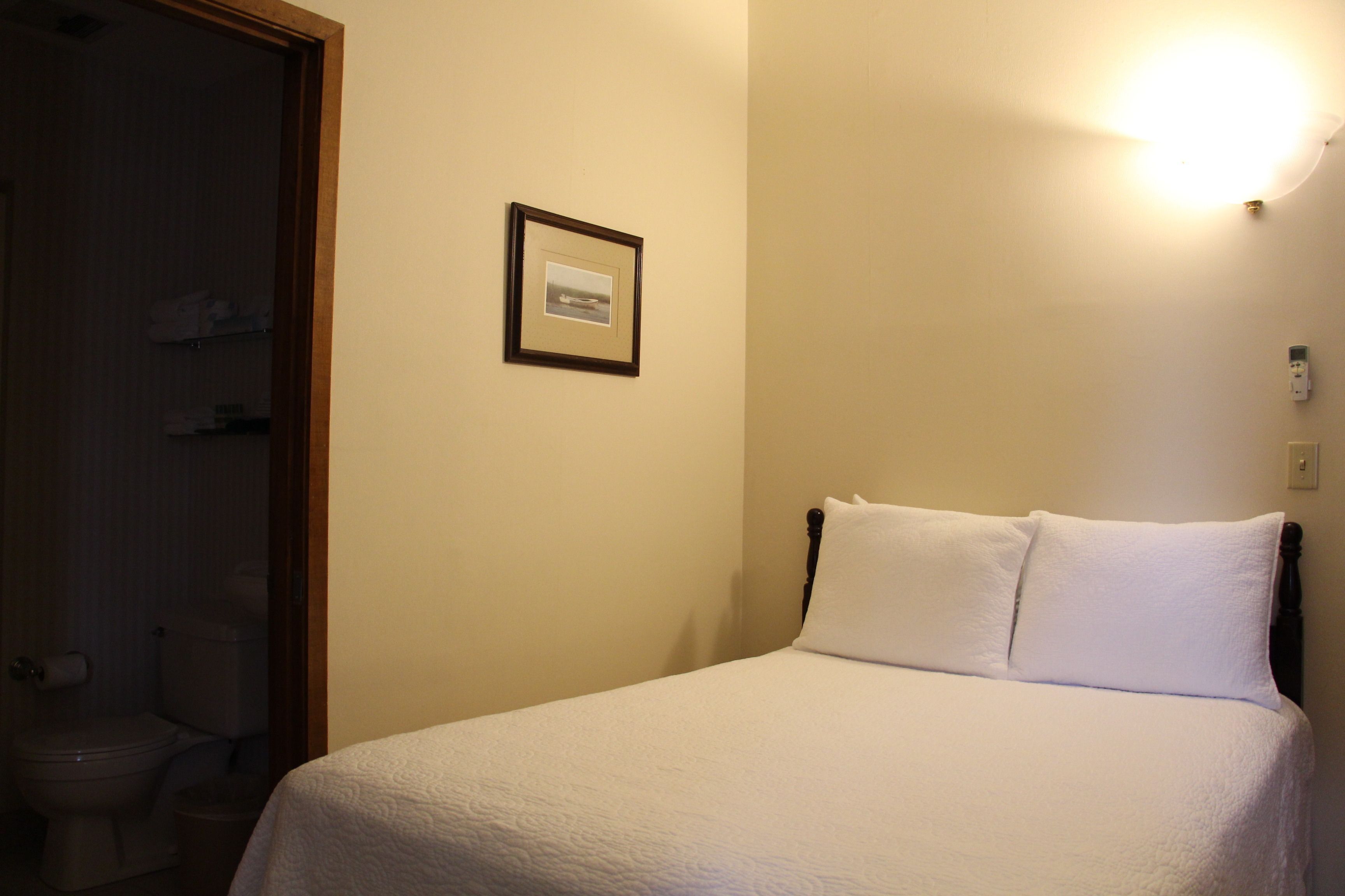 A neatly made bed with white linens in a room with a wooden headboard. A framed picture hangs on the wall above the bed.