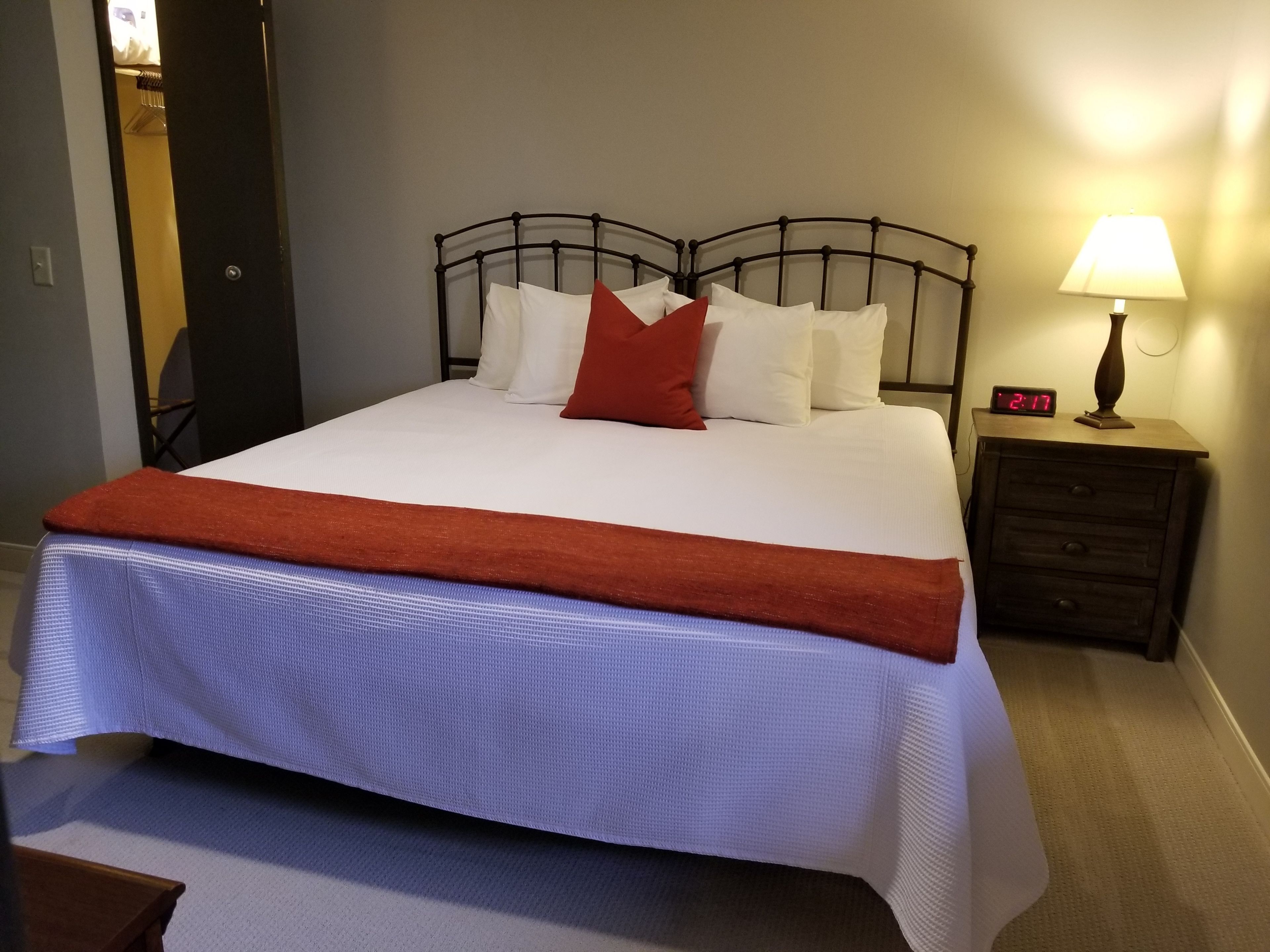 Een net opgemaakt bed met wit en rood beddengoed, een rode plaid en een digitale klok op een nachtkastje.