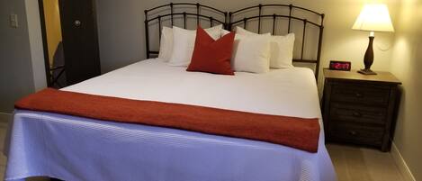 Een net opgemaakt bed met wit en rood beddengoed, een rode plaid en een digitale klok op een nachtkastje.