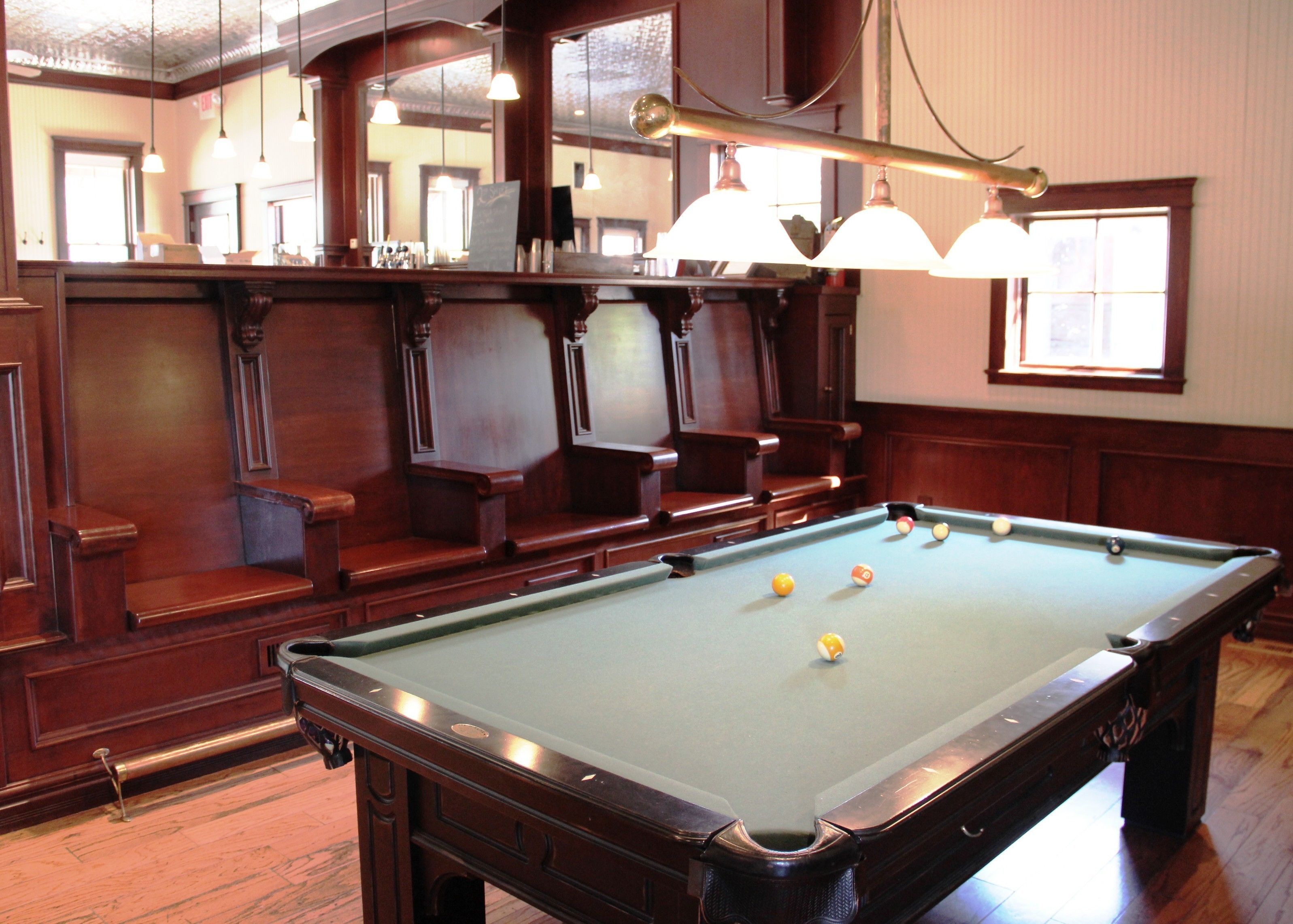 A pool table with balls, wooden benches, and hanging lights in a room with wooden paneling and a window.