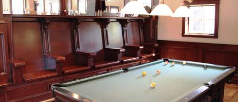 A pool table with balls, wooden benches, and hanging lights in a room with wooden paneling and a window.