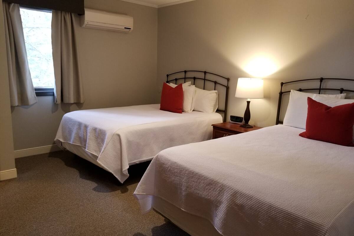 Quarto de hotel com duas camas, mesa de cabeceira com luminária, ar condicionado e janela com cortinas.