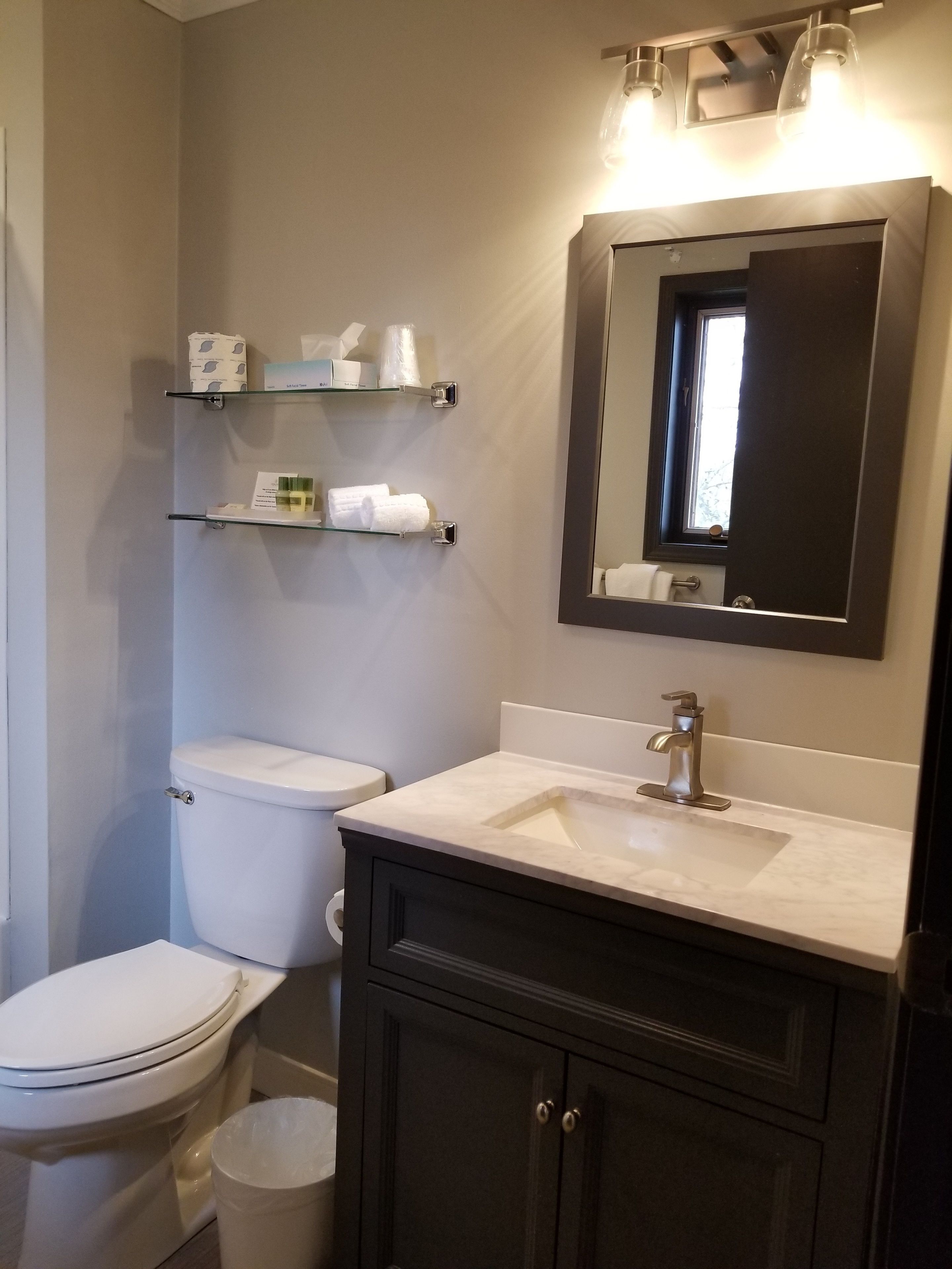 A bathroom with a toilet, a sink, a mirror, and shelves with toiletries.