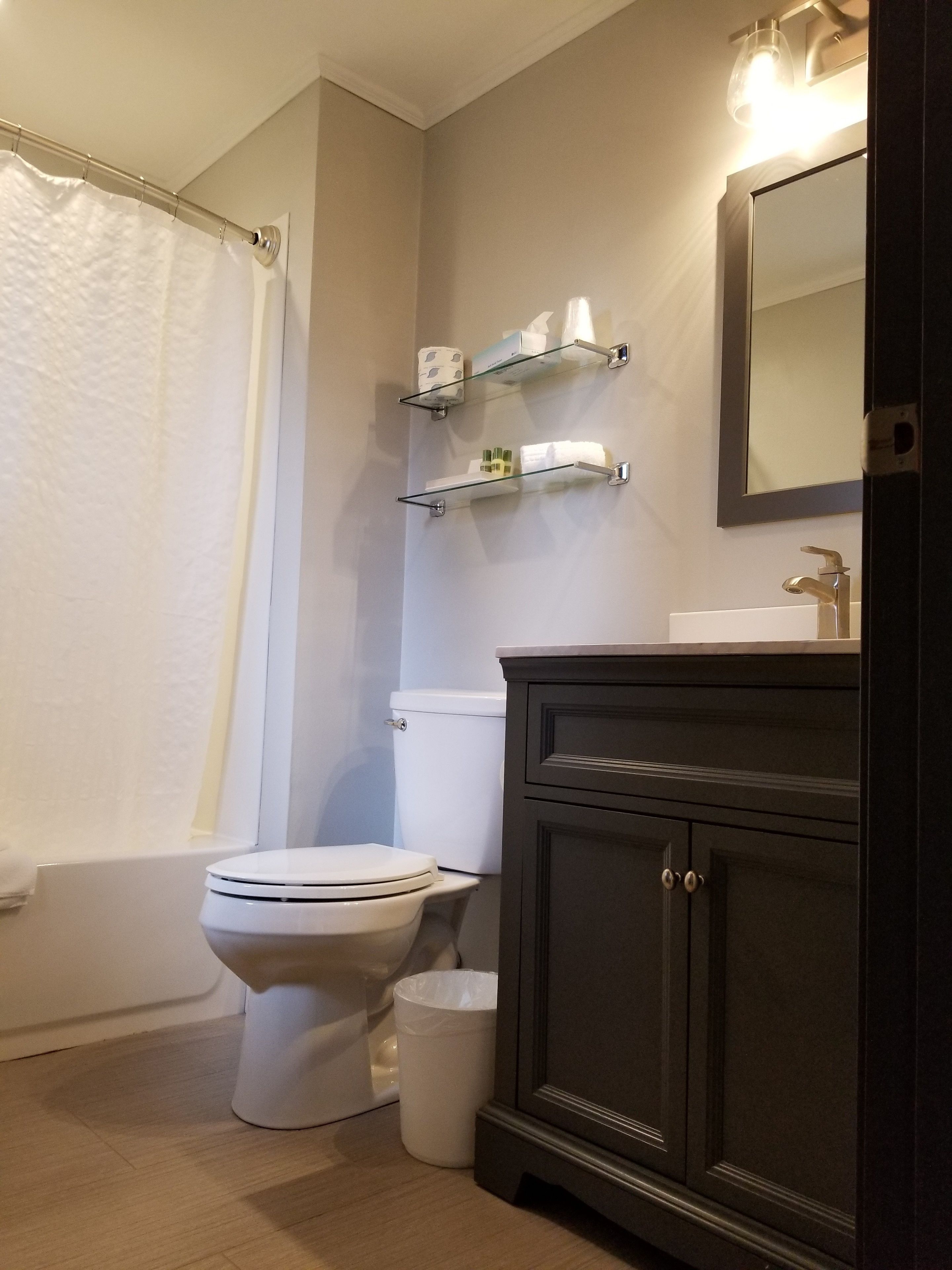 Un baño con inodoro, estante de vidrio con artículos de aseo, espejo y cortina de ducha.