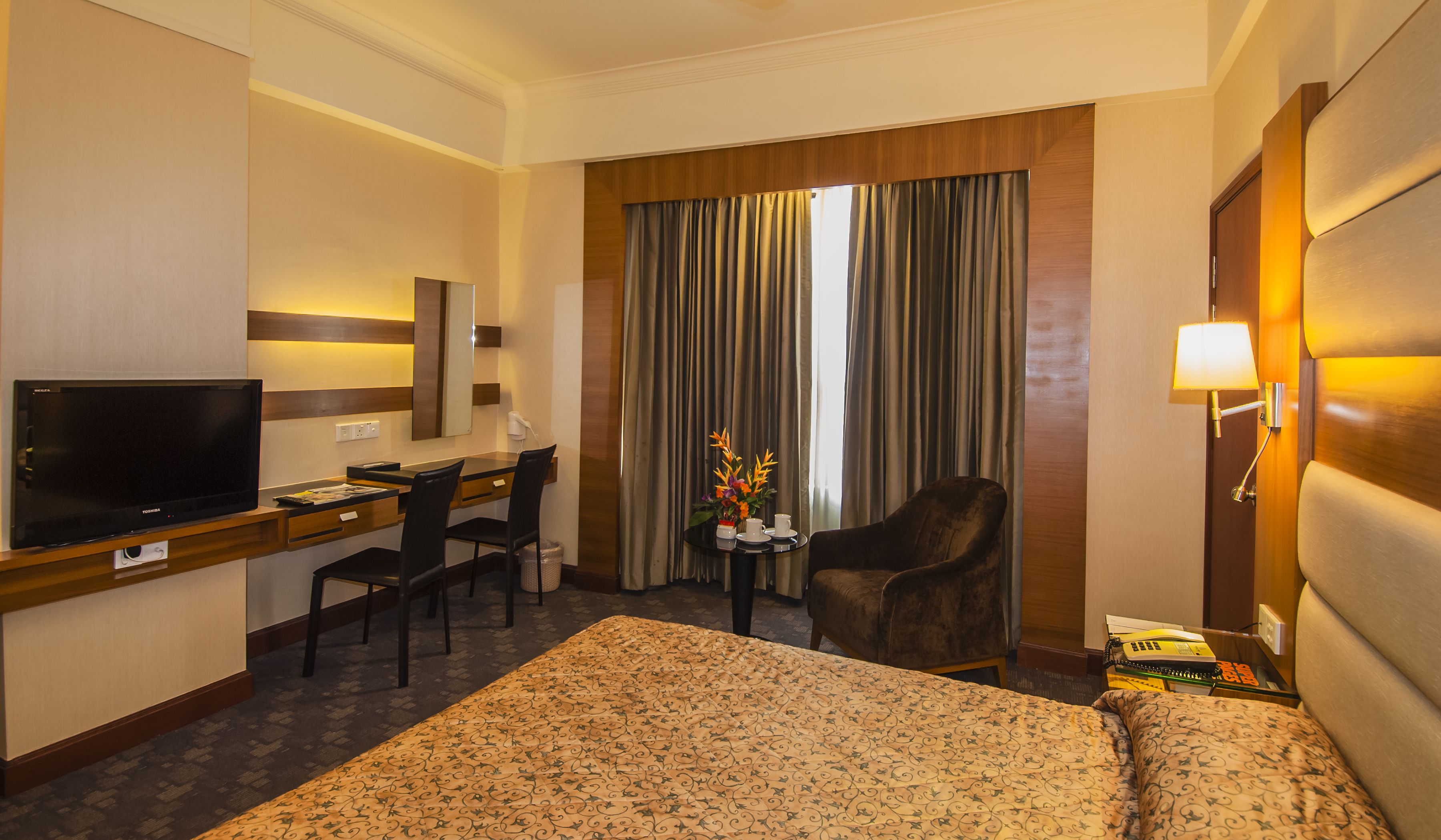 Quarto de hotel com uma cama, uma escrivaninha com televisão, uma cadeira, uma pequena mesa com um vaso e cortinas.