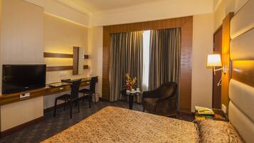 A hotel room with a bed, a desk with a TV, a chair, a small table with a vase, and curtains.
