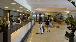 Zona de recepción de un hotel con mostrador de mármol, personal de recepción y una escalera al fondo.