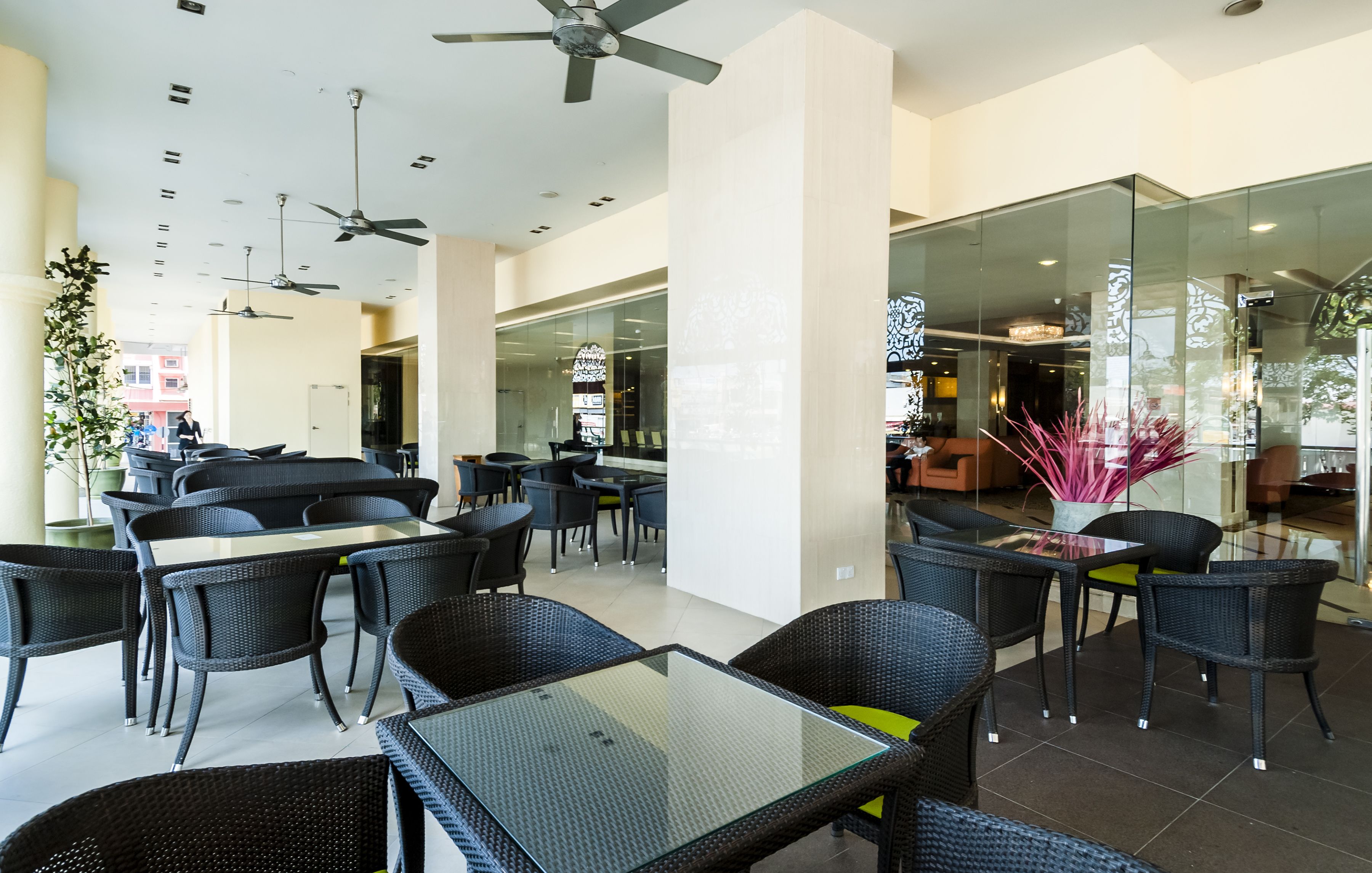 Un espace de restauration intérieur moderne, doté de tables en verre et de chaises en osier, de ventilateurs de plafond et offrant une vue sur la ville grâce à de grandes fenêtres.