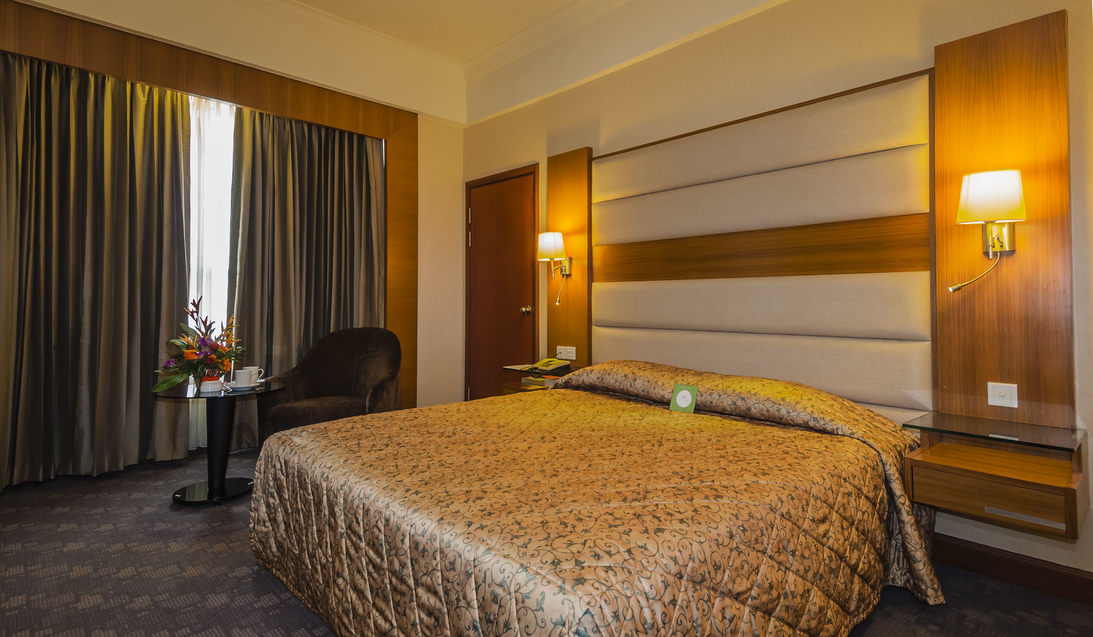 Una habitación de hotel con una cama grande, una silla, una mesita con un florero y una mesita de noche con una lámpara.
