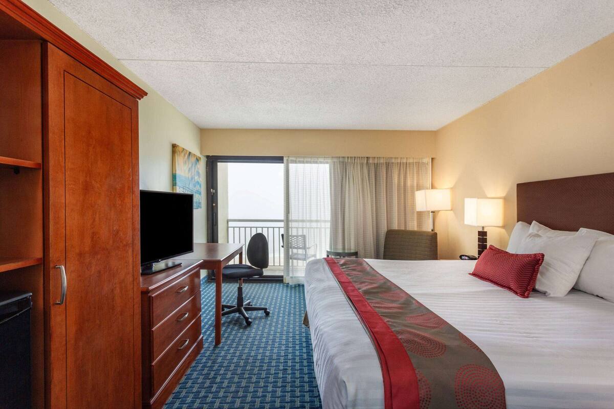 Habitación de hotel con una cama grande, un escritorio con televisor, una silla y un balcón con cortinas.