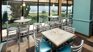 Een eetruimte met meerdere tafels en stoelen, grote ramen met uitzicht op het buitenleven en een muur in teal kleur.