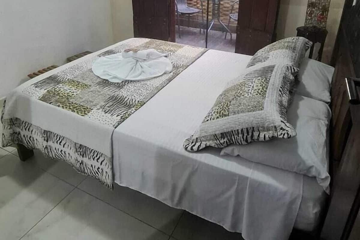 Ein Doppelbett mit gemustertem Bettbezug und Kissen, ein Holfschrank, ein an der Wand befestigter Fernseher und eine Tür zu einem Außenbereich mit Tisch und Stühlen.