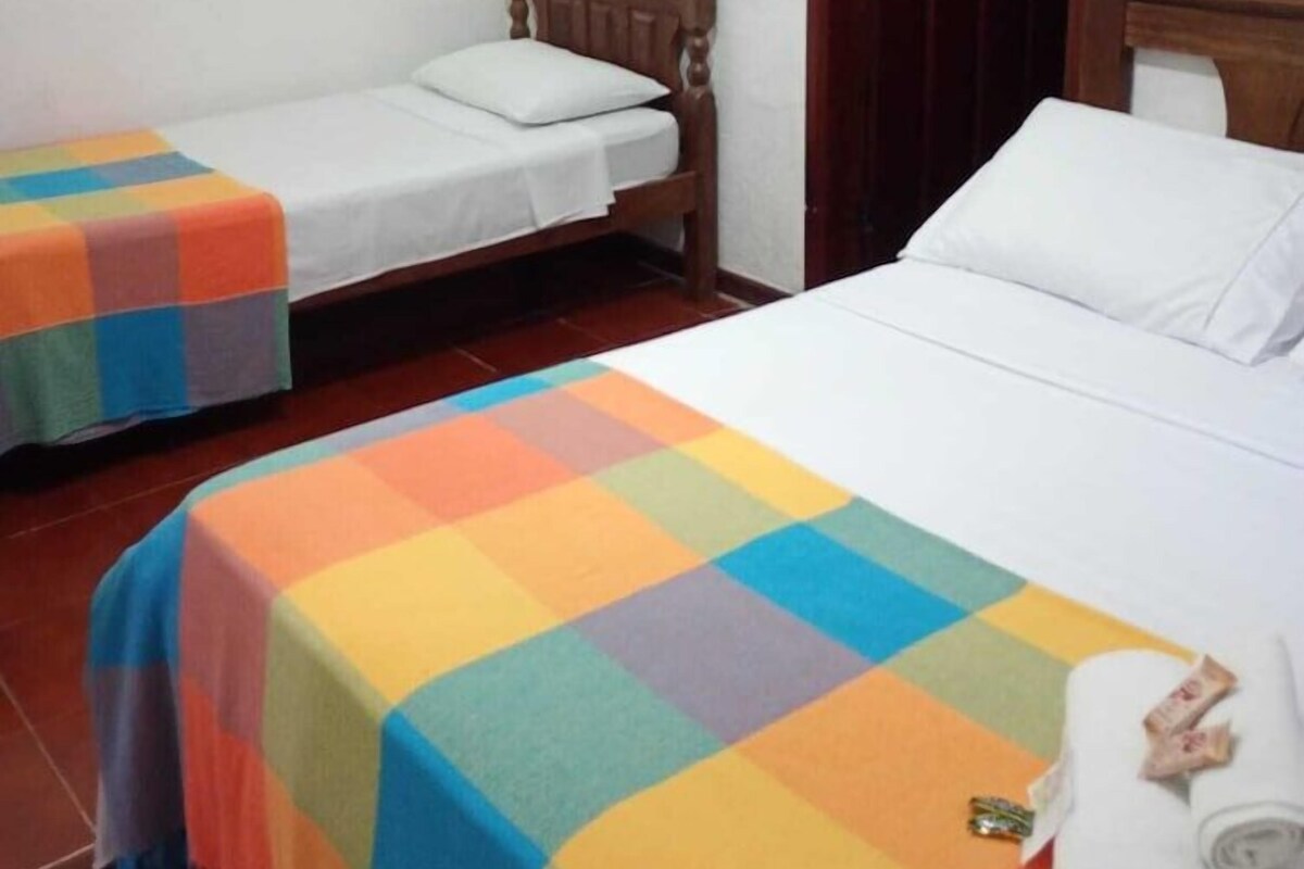 Ein Zimmer mit zwei Betten, eines mit einer farbenfrohen Tagesdecke und das andere mit einer schlichten weißen. Das Bett mit der farbenfrohen Decke hat ein Holz-Kopfteil, und über einer Holztür hängt ein Schild.