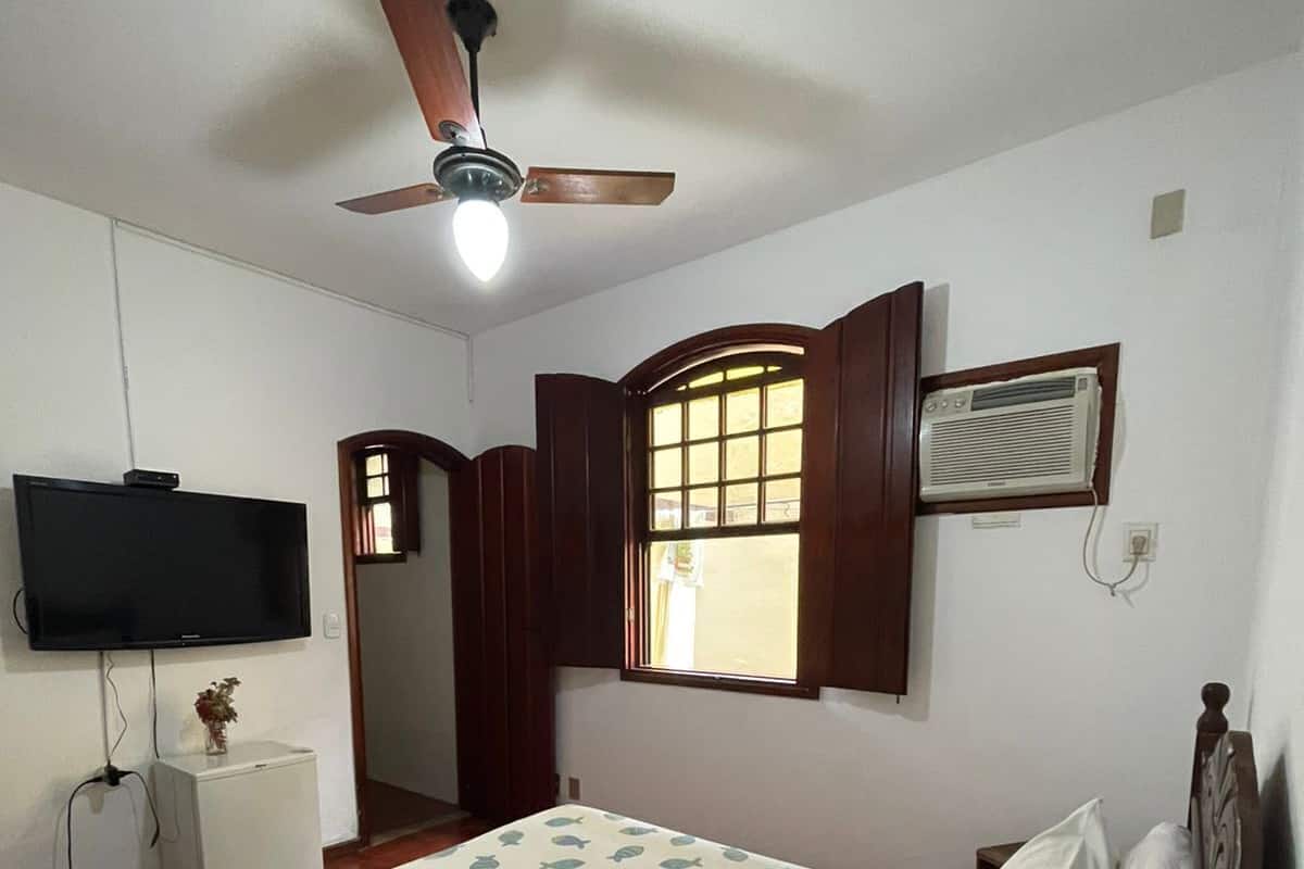 Un dormitorio con cama, ventilador de techo, televisor, ventana con cortinas y aire acondicionado.