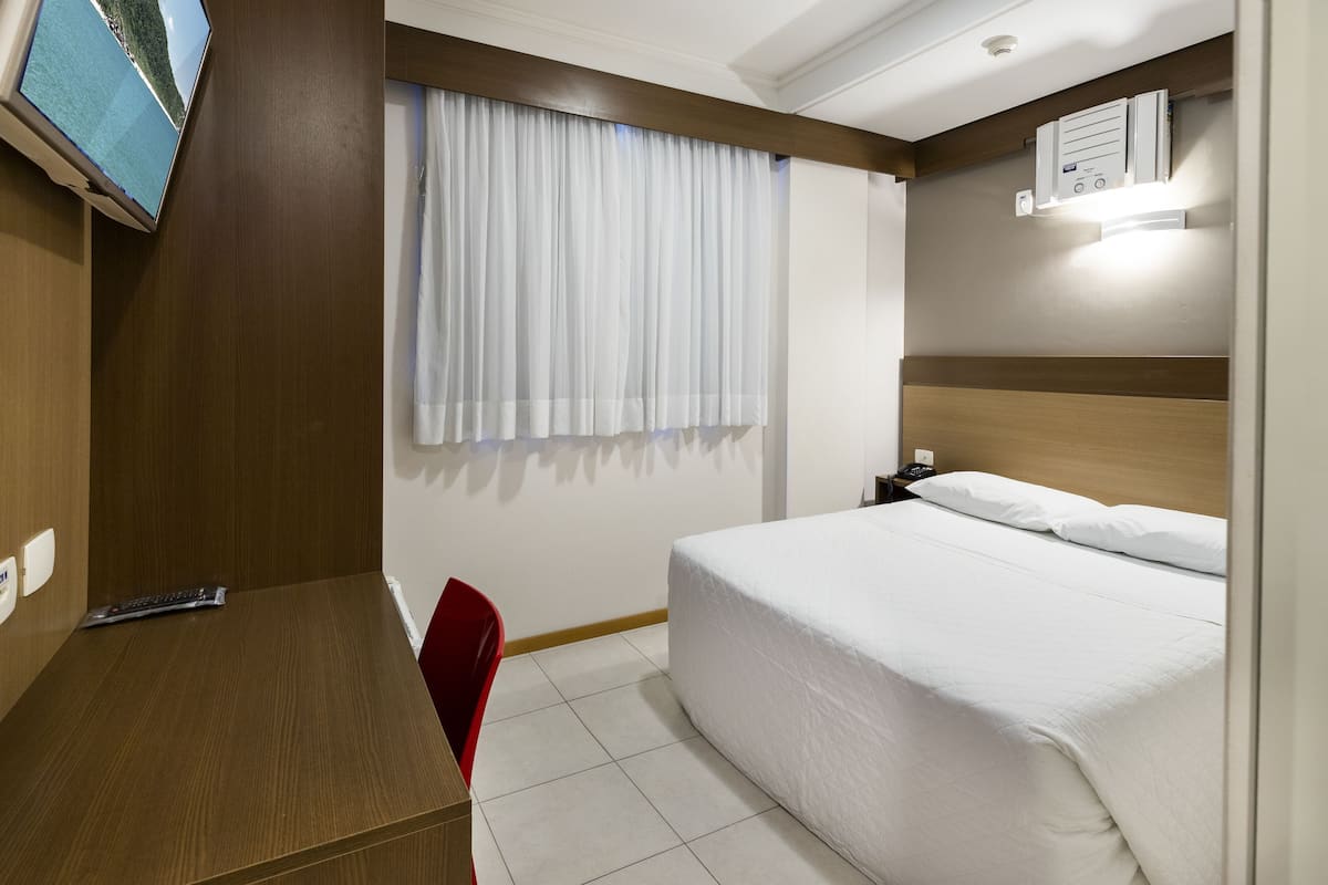 A hotel room with a bed, a desk, a chair, a TV, and a window with curtains.