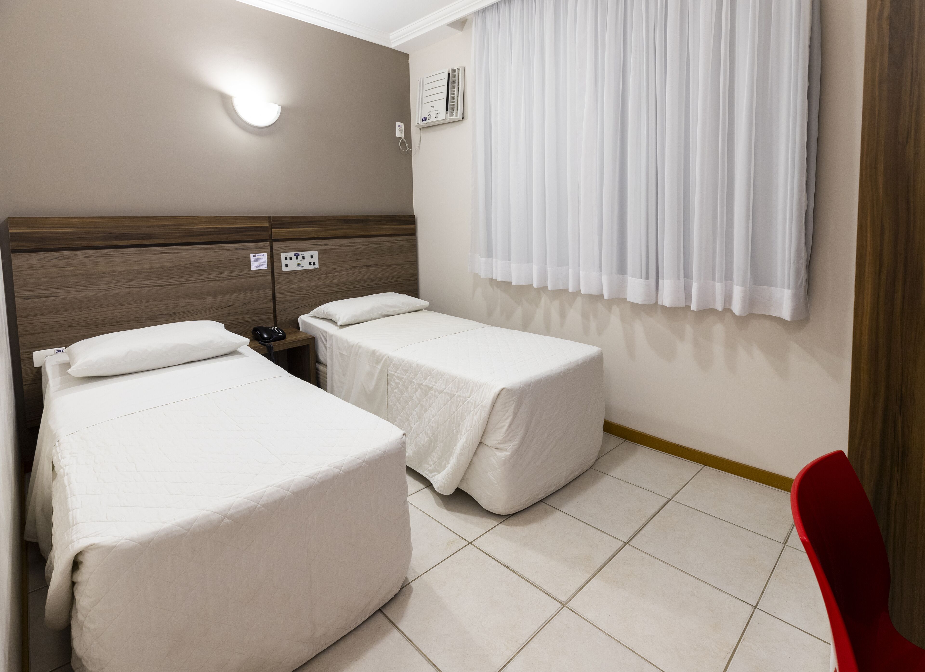 Habitación de hotel con dos camas individuales, cabecera de madera, teléfono junto a la cama, aire acondicionado instalado en la pared y una silla roja.
