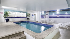 Una piscina interior con baldosas azules cristalinas, una zona de estar con tumbonas blancas y una escalera que conduce a la piscina.