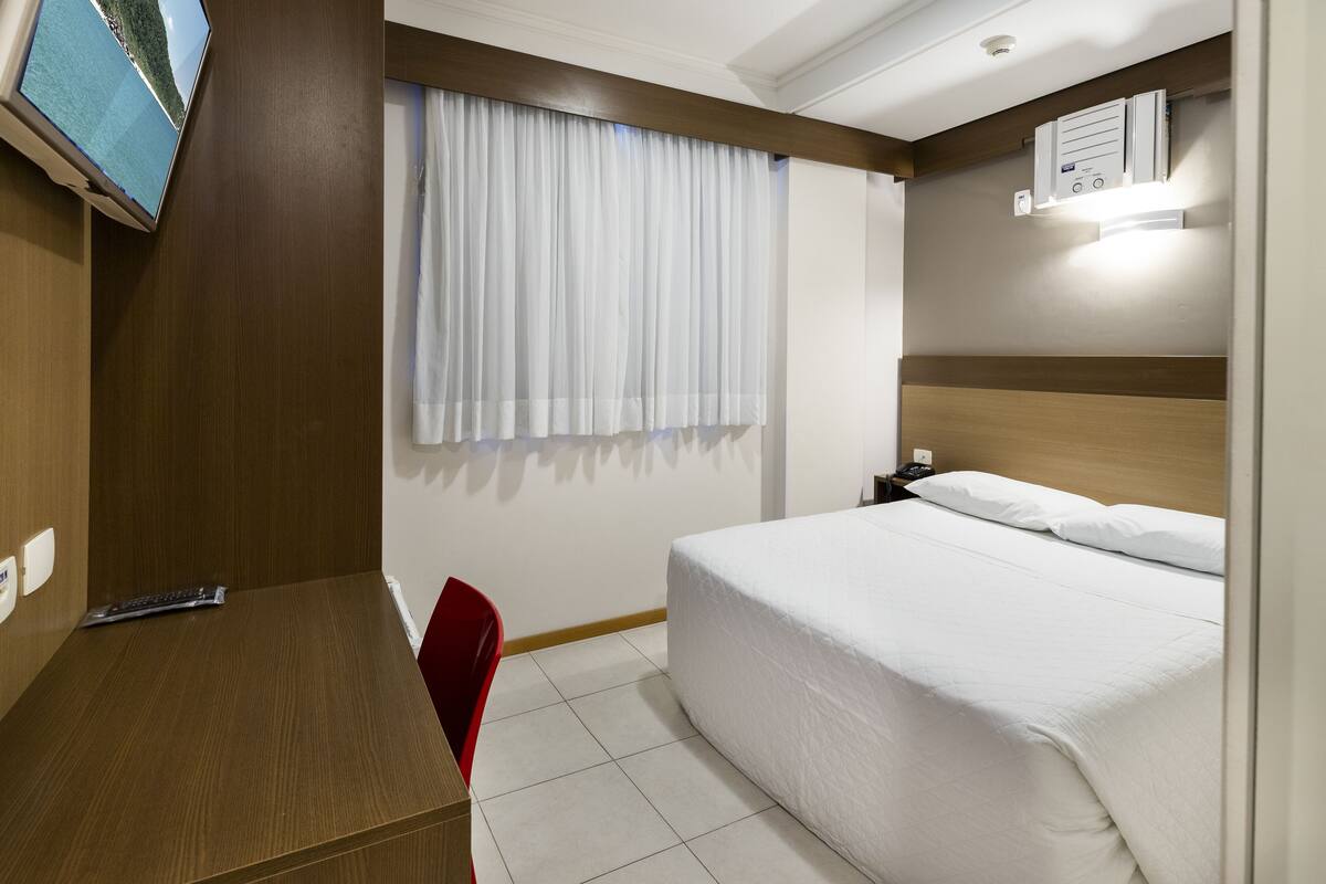 A hotel room with a bed, a desk, a chair, a TV, and a window with curtains.