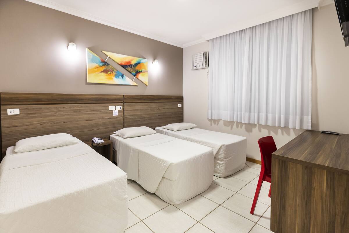 A hotel room with two single beds, a desk, a chair, a painting on the wall, and a window with curtains.