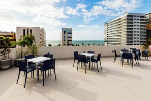 An outdoor terrace with tables and chairs, overlooking a body of water and buildings. - El Aram Beach Express (Maceió)