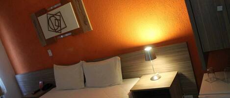 Habitación de hotel con cama, mesita de noche con lámpara, mesita auxiliar y una obra de arte en la pared.