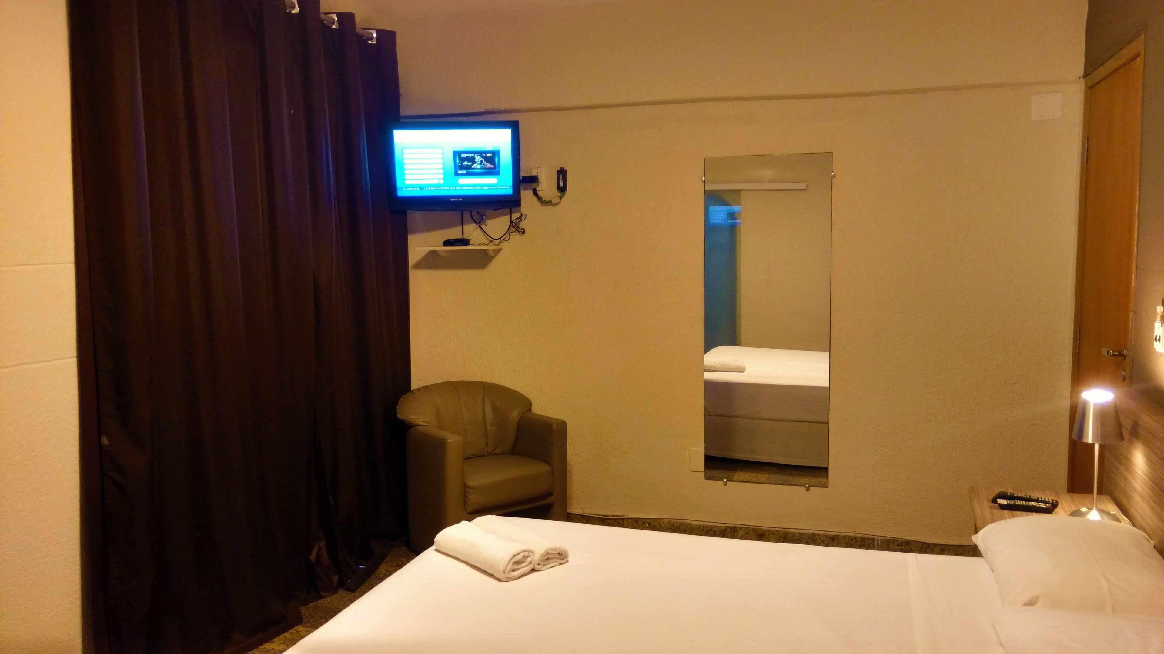 Una habitación de hotel con cama, sillón, televisor, espejo y mesita de noche con control remoto.