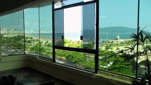 A room with a large window overlooking a cityscape, a view of the beach, and a chair in the foreground. - Hotel Imperador (Santos)