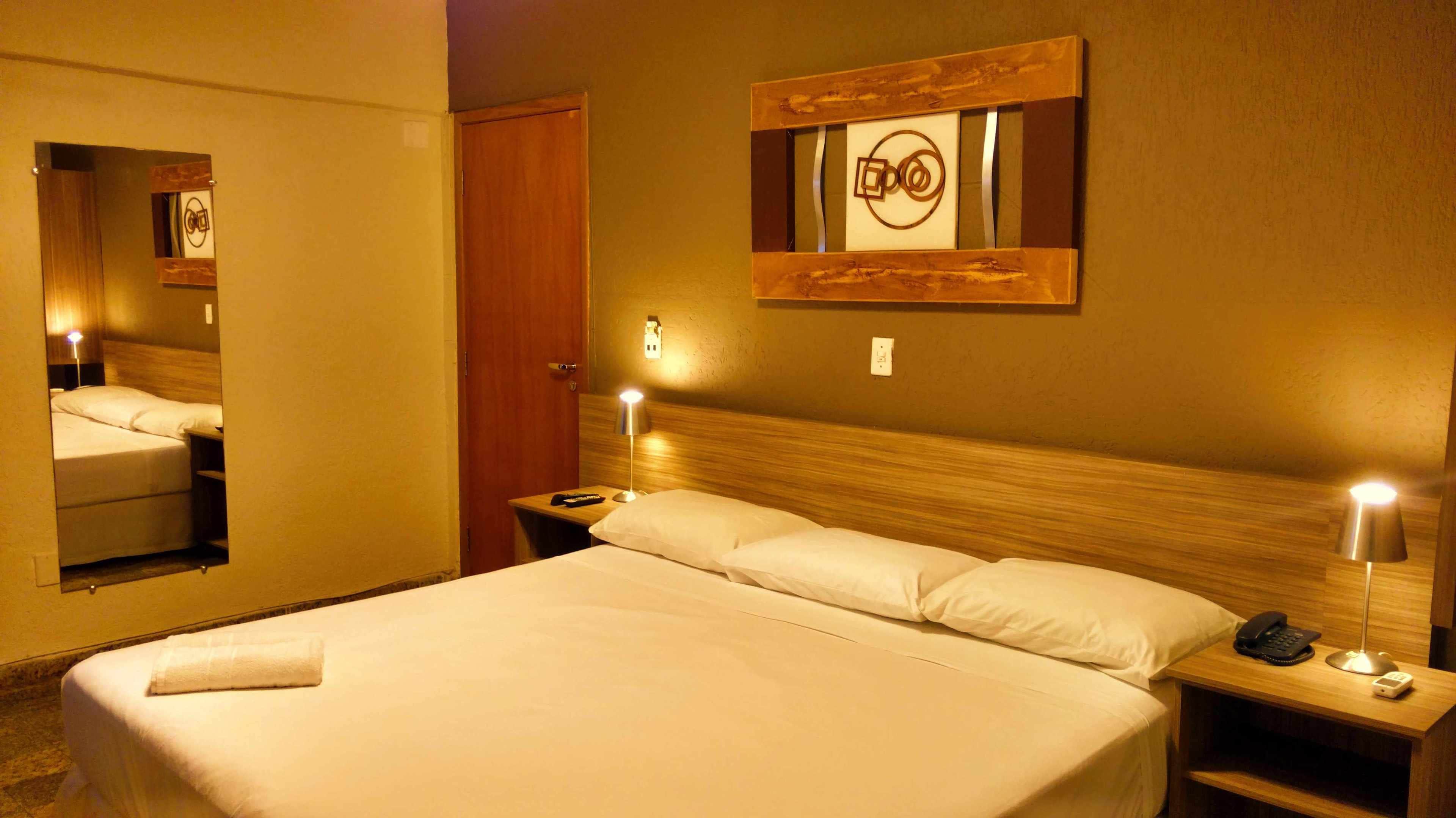 A hotel room with a large bed, bedside tables with lamps, a wooden headboard, and a mirror on the wall.