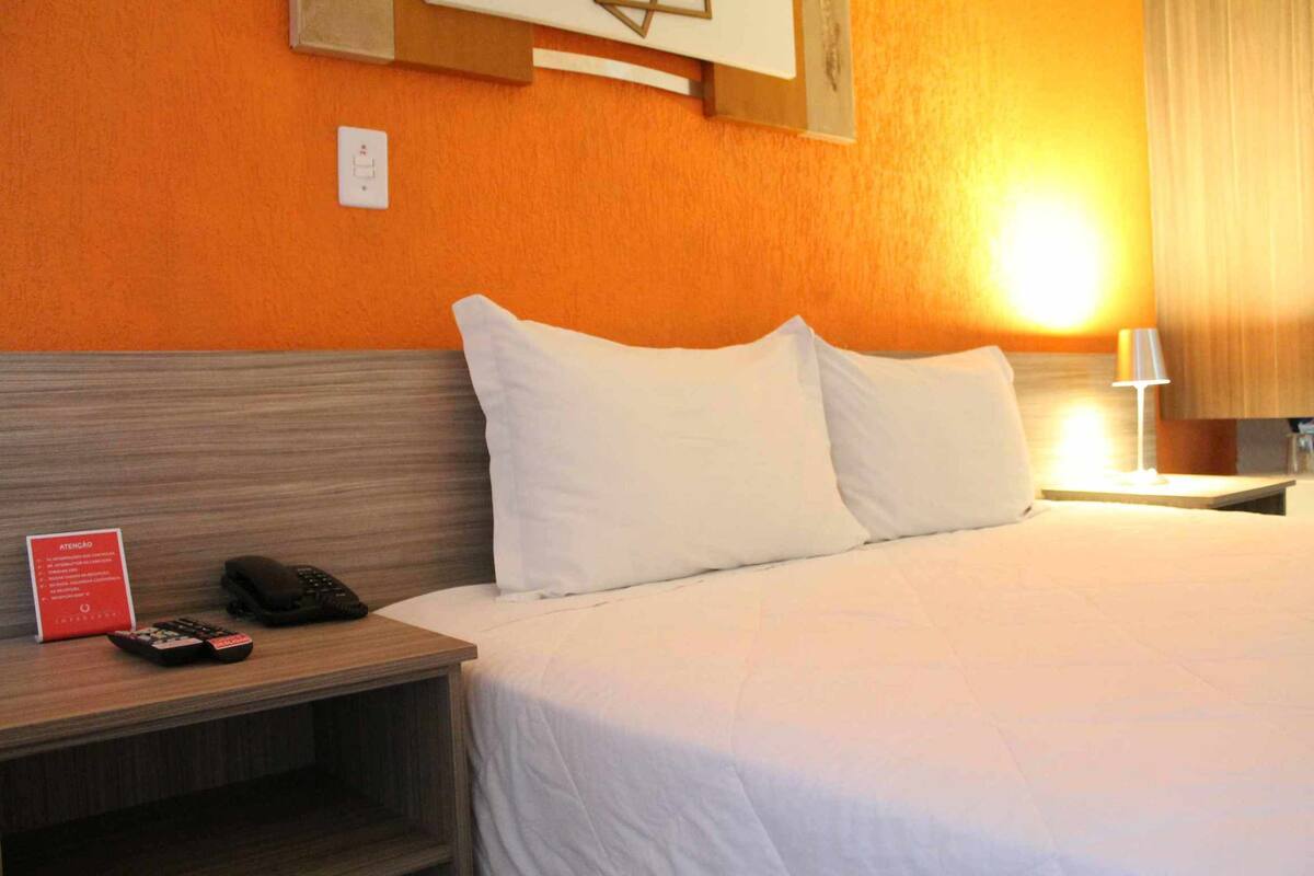 A hotel room with a bed, white pillows, a nightstand with a phone and remote controls, and a sign with instructions.