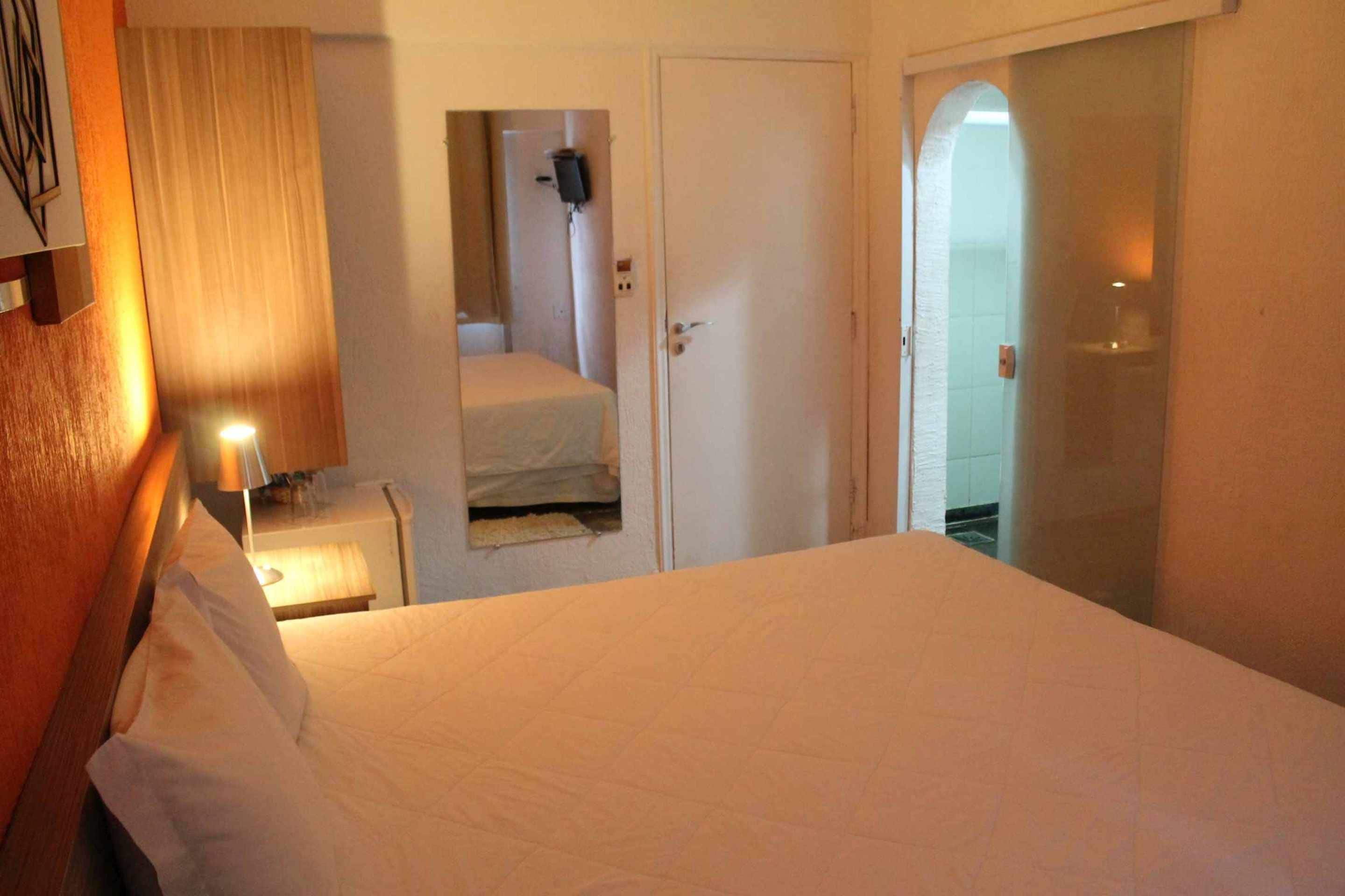 A hotel room with a bed, a bedside table with a lamp, a door leading to a bathroom, and a mirror on the wall.