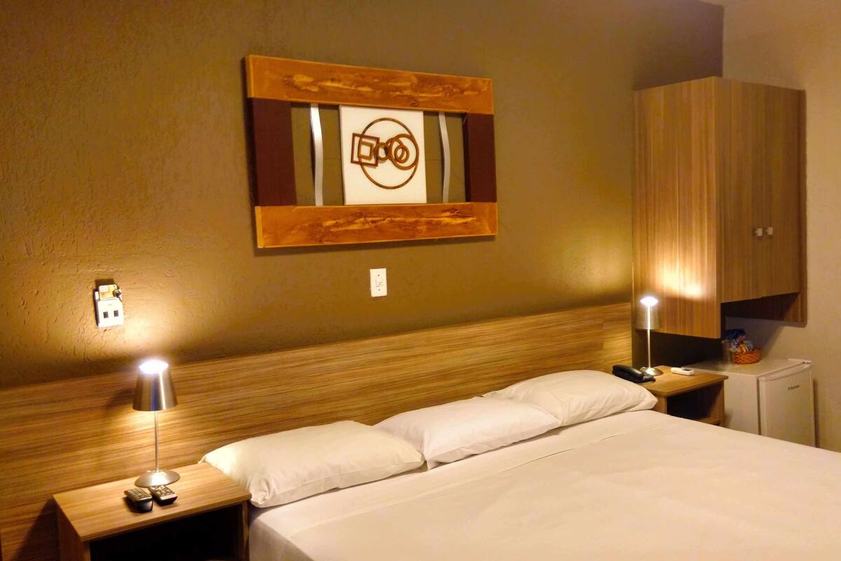 A hotel room with a wooden headboard, a bed with white linens, two bedside lamps, a nightstand with a remote control, and a wall-mounted artwork.