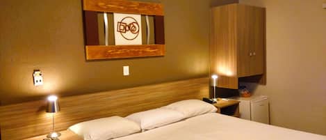 Hotelkamer met een houten hoofdeinde, een bed met witte lakens, twee bedlampjes, een nachtkastje met een afstandsbediening en een kunstwerk aan de muur.