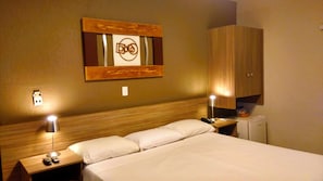 A hotel room with a wooden headboard, a bed with white linens, two bedside lamps, a nightstand with a remote control, and a wall-mounted artwork. - Hotel Imperador (Santos)