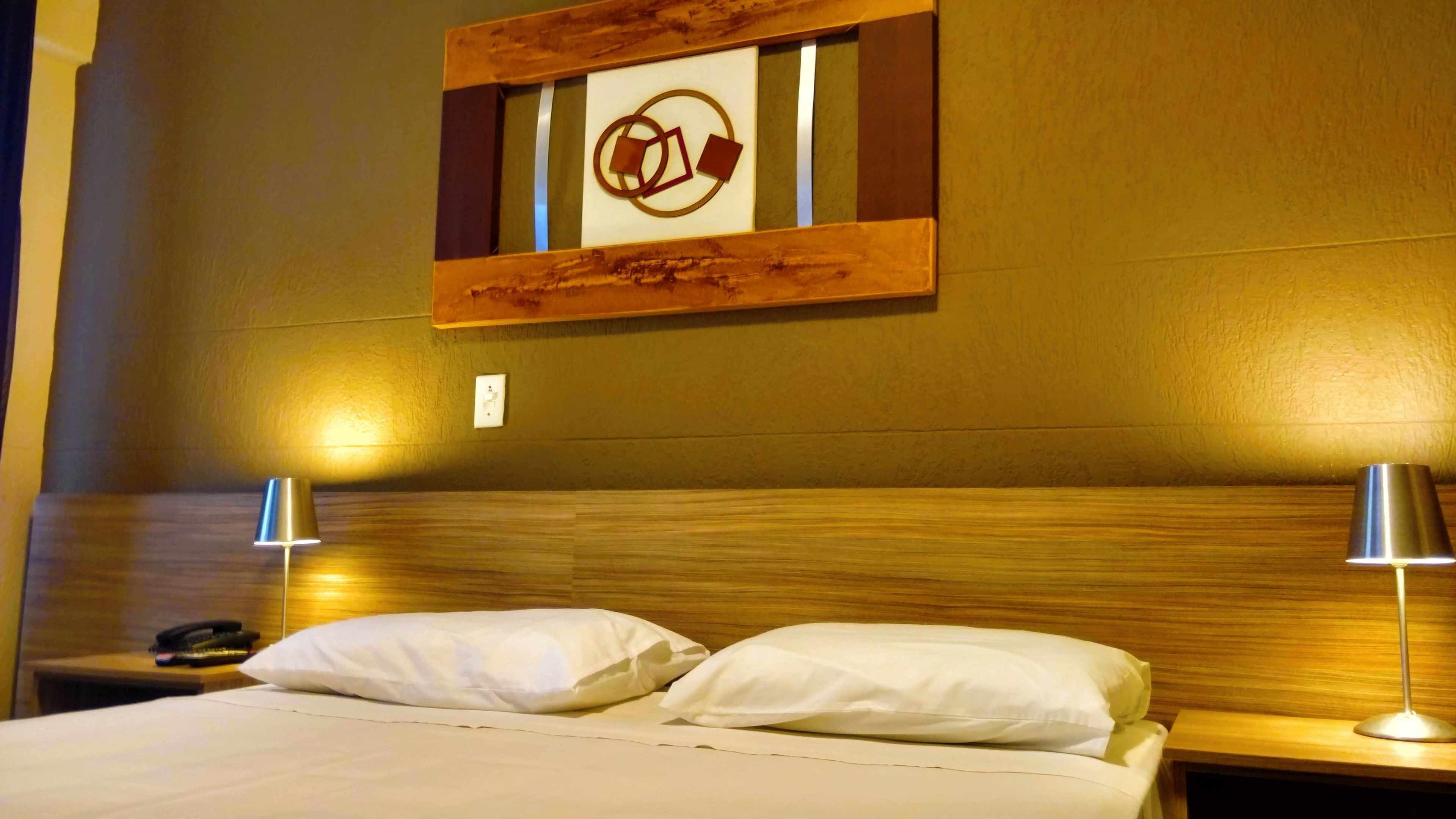 Habitación de hotel con cabecera de madera, dos lámparas de noche, cama con ropa blanca y una obra de arte colgada en la pared.