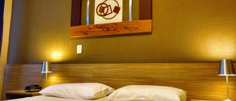Habitación de hotel con cabecera de madera, dos lámparas de noche, cama con ropa blanca y una obra de arte colgada en la pared.