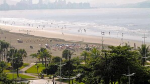 A beach with umbrellas and people, palm trees, and a city skyline in the background. - Hotel Imperador (Santos)